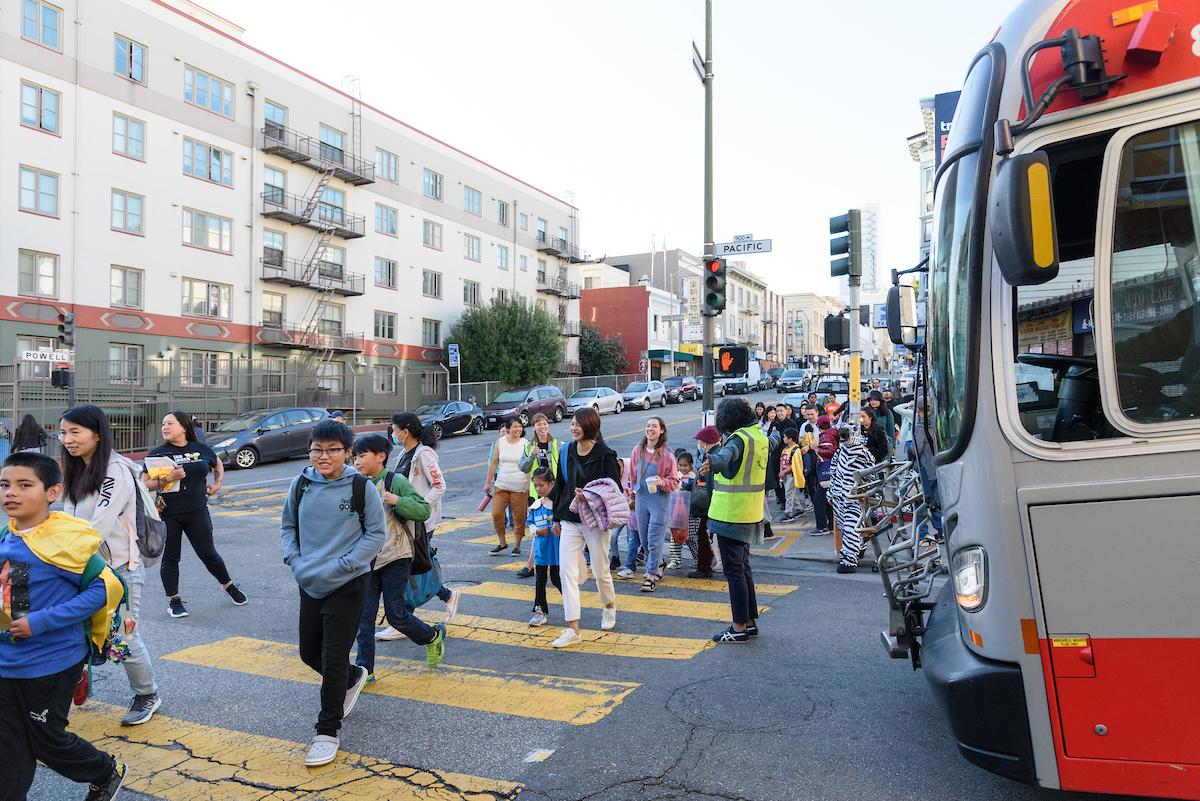 Students and families cross a street with help from a crossing guard. A bus pauses to let everyone through the intersecion.