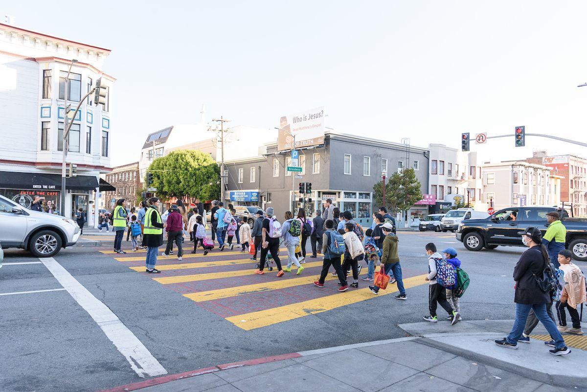 Students and families cross an intersection with help from crossing guards wearing yellow safety vests.