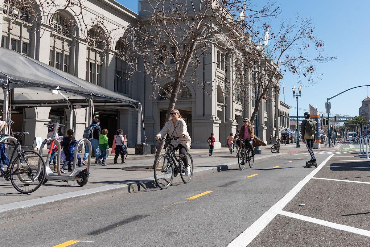 People walk on the sidewalk and others bike on the nearby street.