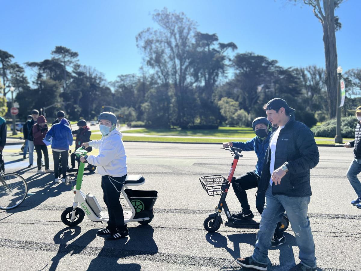 Two young people use adaptive scooters in Golden Gate Park while others walk on the car-free roadway and stand nearby.