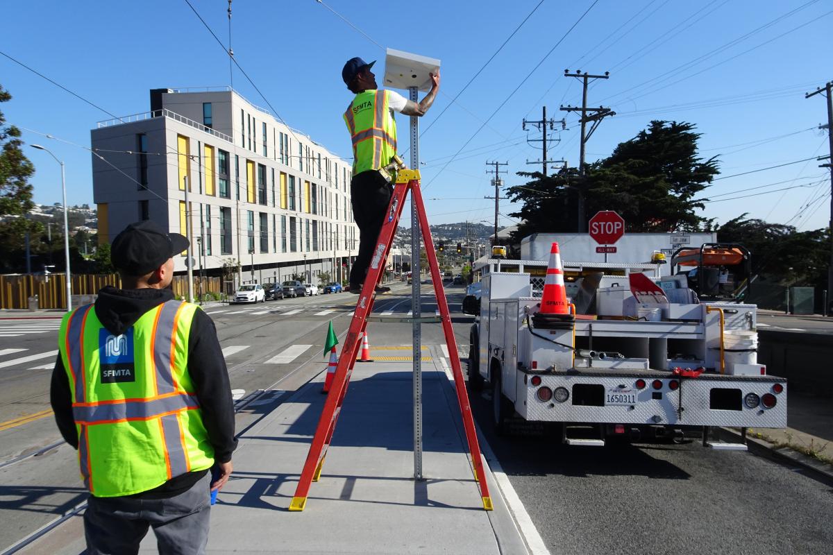 Photo of two SFMTA staff on a boarding platform. One is on a ladder installing a solar panel on top of a pole while another observes.