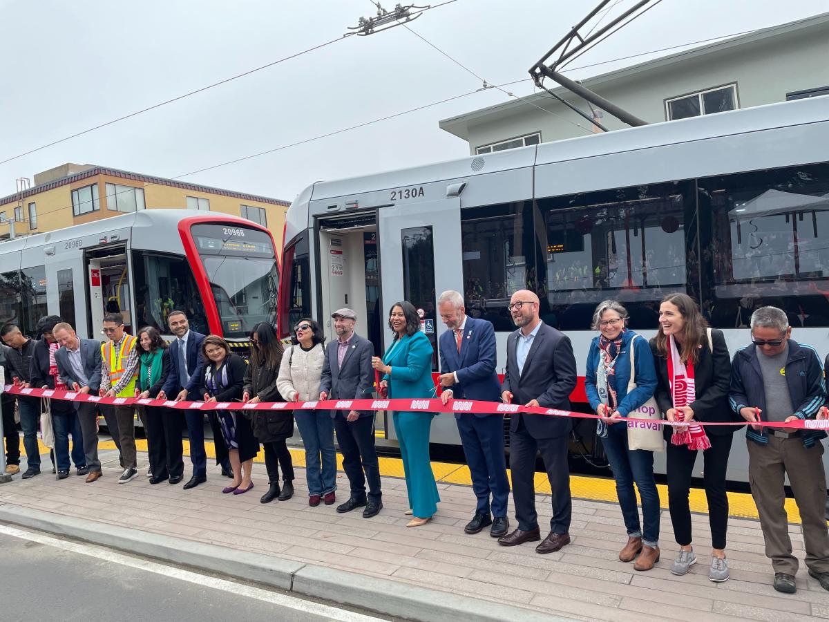 Several people holding a large red ribbon stand in front of an L Taraval train.