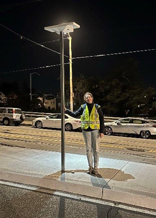 SFMTA staff person stands under the lit pilot solar light installed on a boarding platform. 