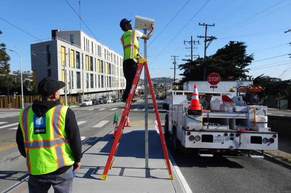 Photo of two SFMTA staff on a boarding platform. One is on a ladder installing a solar panel on top of a pole while another observes.