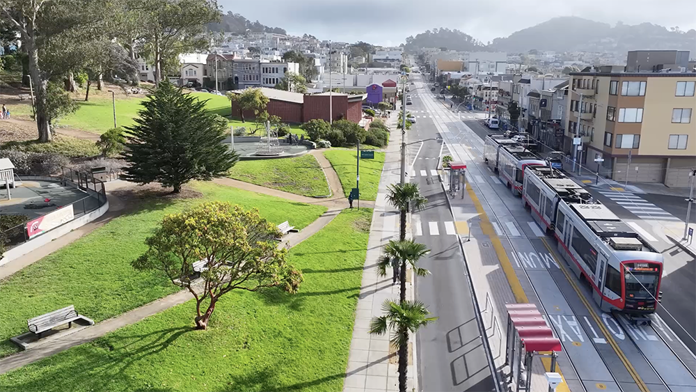 Aerial image of an L Taraval train moving down the street, passing a green space with trees and pathways.