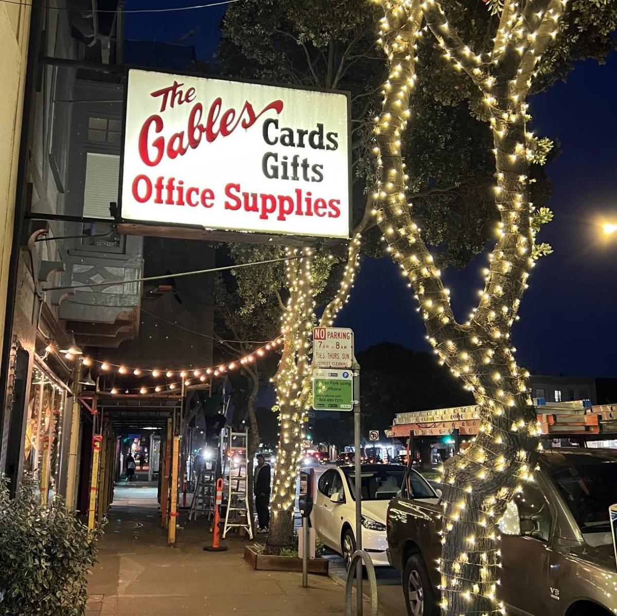 Image shows multiple trees with decorative lighting near the Gables Office Supplies sign on Geary Boulevard.