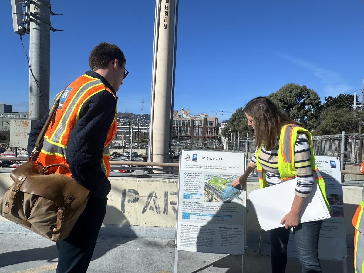 a man in an orange safety vest and woman in a yellow safety vest look at a board with information