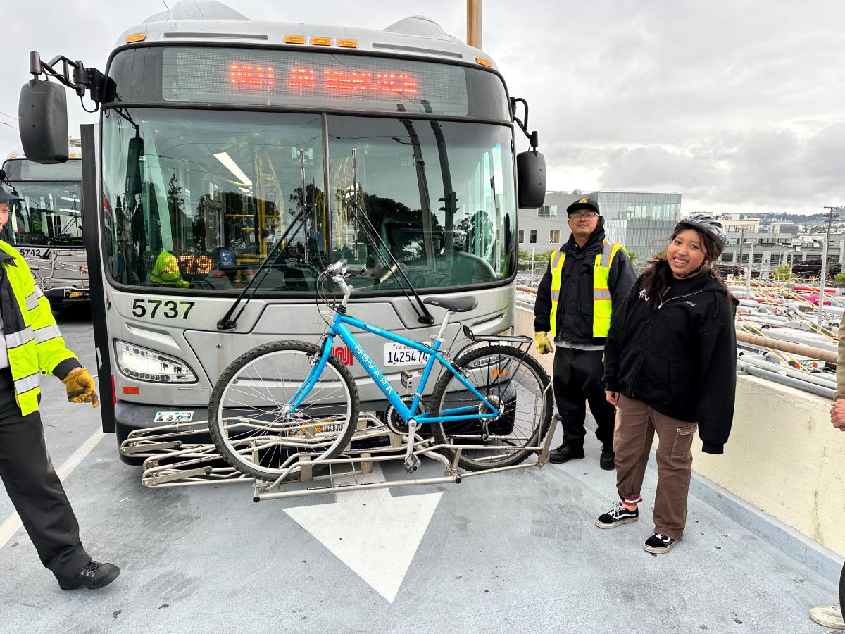 Two people smile in front of a Muni bus that has a bike on the rack.