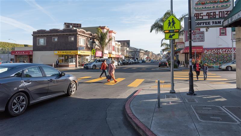 Two people cross a street on a commercial corridor as a car waits.