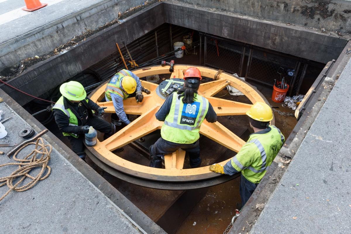 Several people wearing yellow SFMTA vests reinstall a cable car sheave below the street's surface.