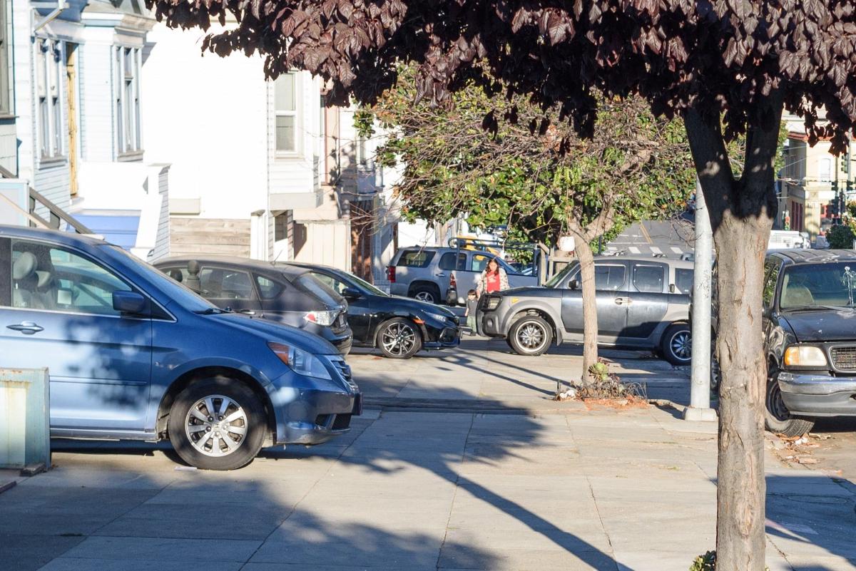 Image shows several vehicles parked illegally over the sidewalk on a hilly street with a parent and child walking around them as they climb the hill.