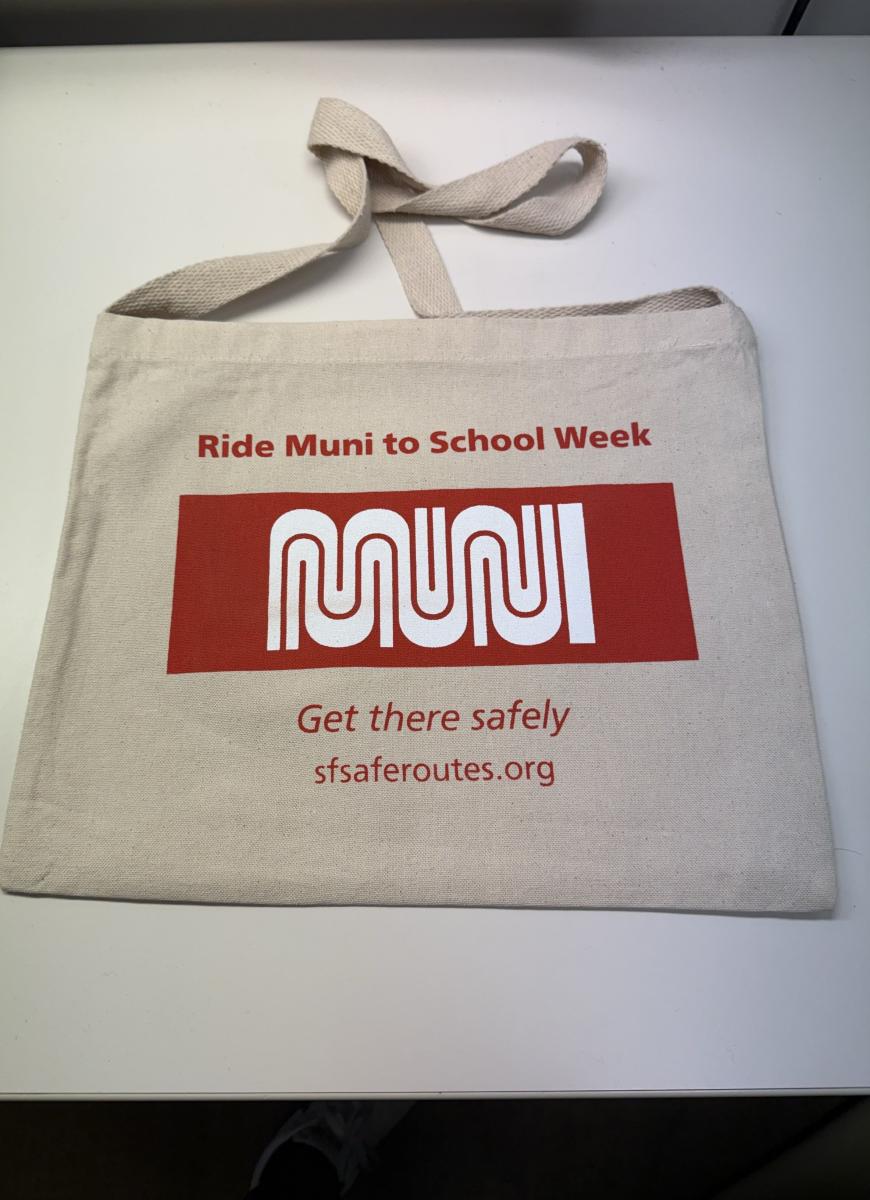 Image of a beige and red Ride Muni to School Week tote bag placed on a desk.