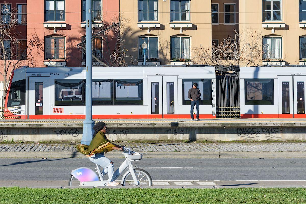 Woman rides a bikeshare bike on a street near a grassy area as a man waits on a train platform in the background.