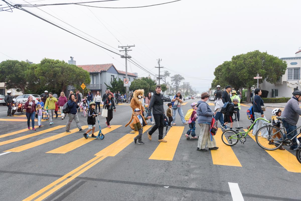 Students, caregivers and others walk and bike in a large group through a yellow crosswalk.