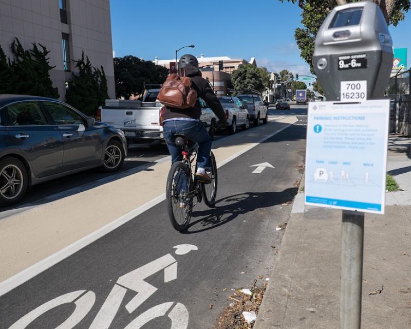 A person bikes on Valencia Street in a bike lane placed between a lane of parked cars and a sidewalk curb, with parking meters featuring instructional flyers.