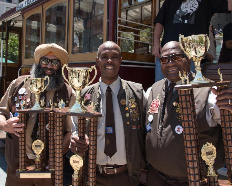 Three Muni operators stand with trophies in front of a motorized cable car in Union Square.