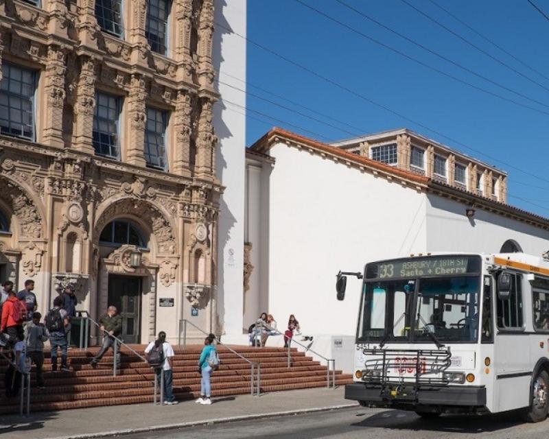 White and orange Muni trolley bus sits in front of Mission High School while students mingle on the front steps.
