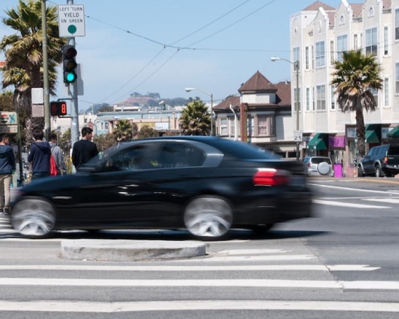 A car appears blurry as it moves through a crosswalk in a San Francisco intersection with pedestrians crossing in the background.