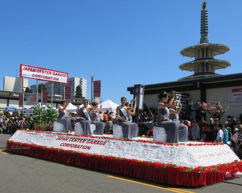 Ladies in gray dresses sit on a red and white float waving to the crowd under a bright blue sky.