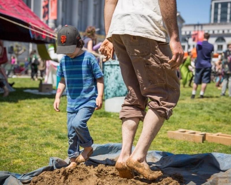 A man and child walk barefoot in mud at the Earth Day Festival in Civic Center Plaza.