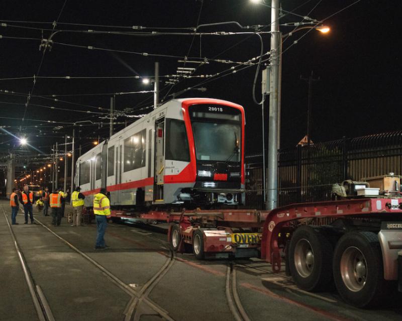 A red and gray light rail vehicle is off-loaded from a flat-bed truck at night with crew members in safety gear standing by. 