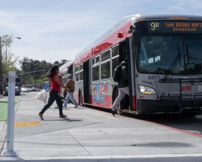 People board a Muni bus at a stop using a new bus boarding island, with a bike lane placed between the island and the sidewalk, on the 9R San Bruno Rapid route.