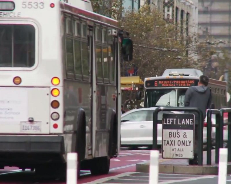 Muni buses on Market Street travel down red-colored transit-only lanes, approaching riders waiting on a boarding island, with a bus shelter labeled, "Muni Rapid."