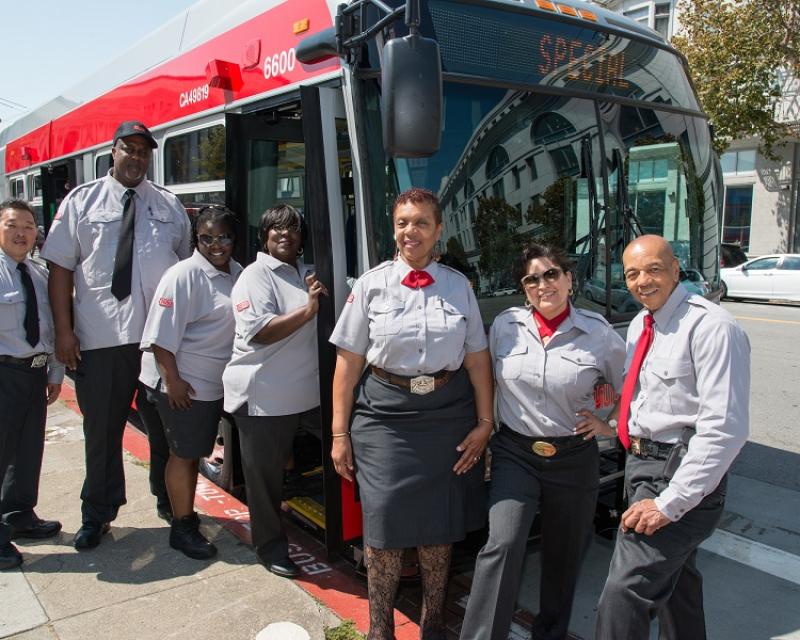 Muni operators pose in their new uniforms in front of a parked Muni bus.