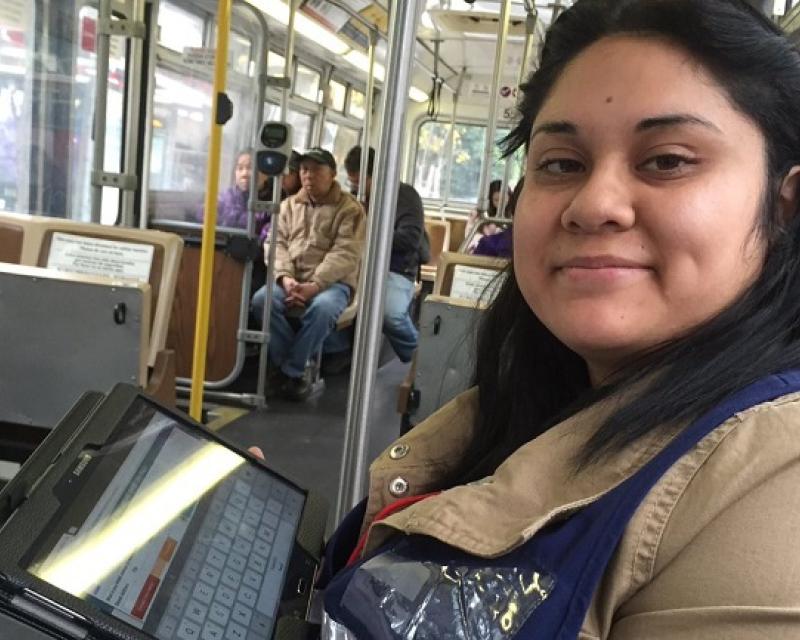 An image of a Muni surveyor in a blue vest and beige coat sitting on a Muni bus holding a black tablet.
