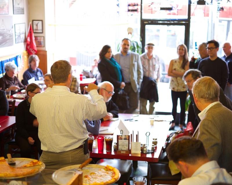 Meeting attendees listen to a speaker in a small pizzeria.