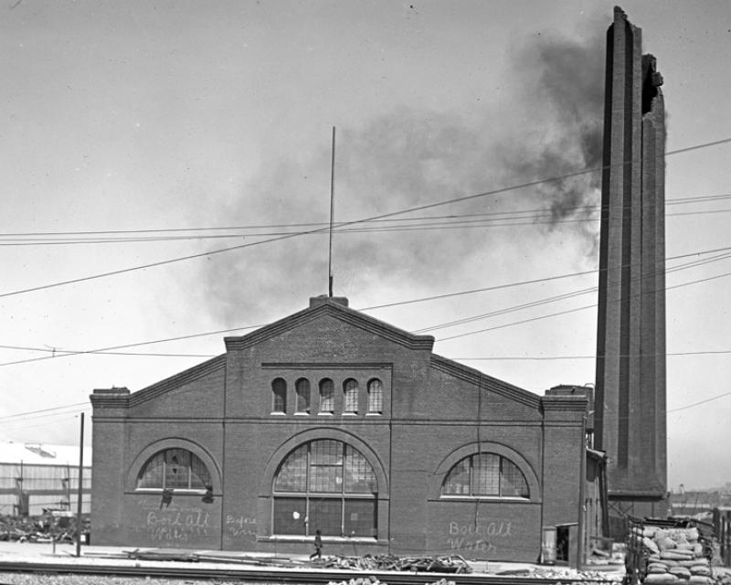 Black and white photo taken in June, 1906 of a large brick building on Market and Valencia Streets. The building is partially damaged with the top of a tall smokestack broken off and light black smoke coming out of it.  Surrounding the building are piles of rubble from destroyed and damaged buildings.