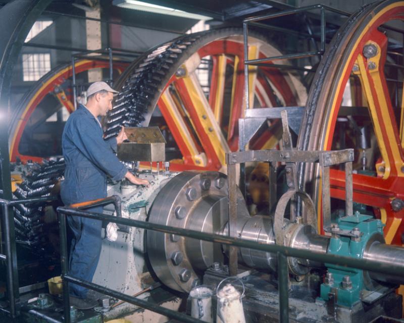 Color photo of a man standing next to the huge pulleys of the cable winding machinery, looking into a bronze oil box on top of a large bearing housing.