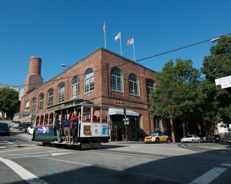 A crowded cable car traveling past the Cable Car Museum and Powerhouse on a sunny day.