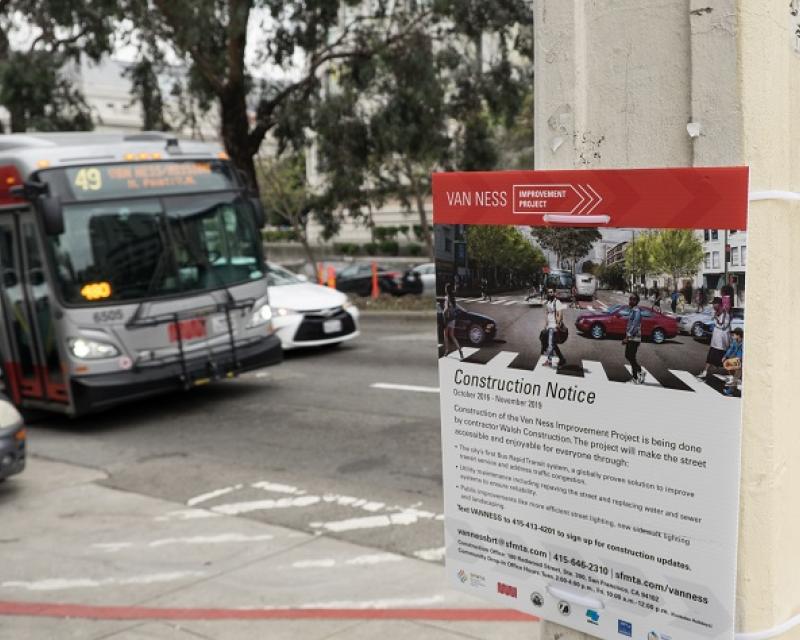 Bus driving up Van Ness Avenue, foreground is a construction notice posted for the Van Ness Improvement Project.