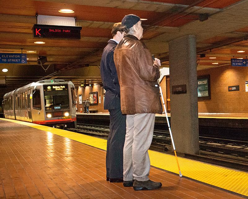 Two passengers waiting for Muni Metro on the platform at Castro Station, one passenger uses a walking cane as a mobility tool