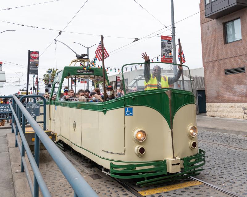 Cream and green open-air streetcar, a Boat Tram, drives near the Embarcadero. The operator waves.