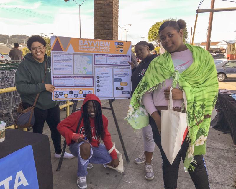 Students smile around a sign featuring information about the Bayview Community Based Transportation Plan. 