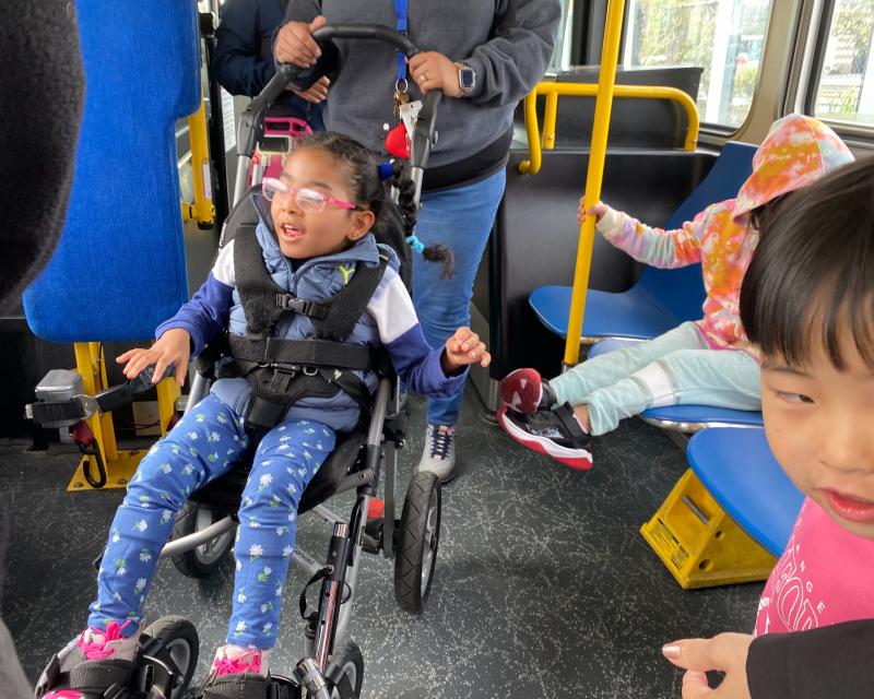 A passenger in a mobility stroller smiles near other young children on a bus.