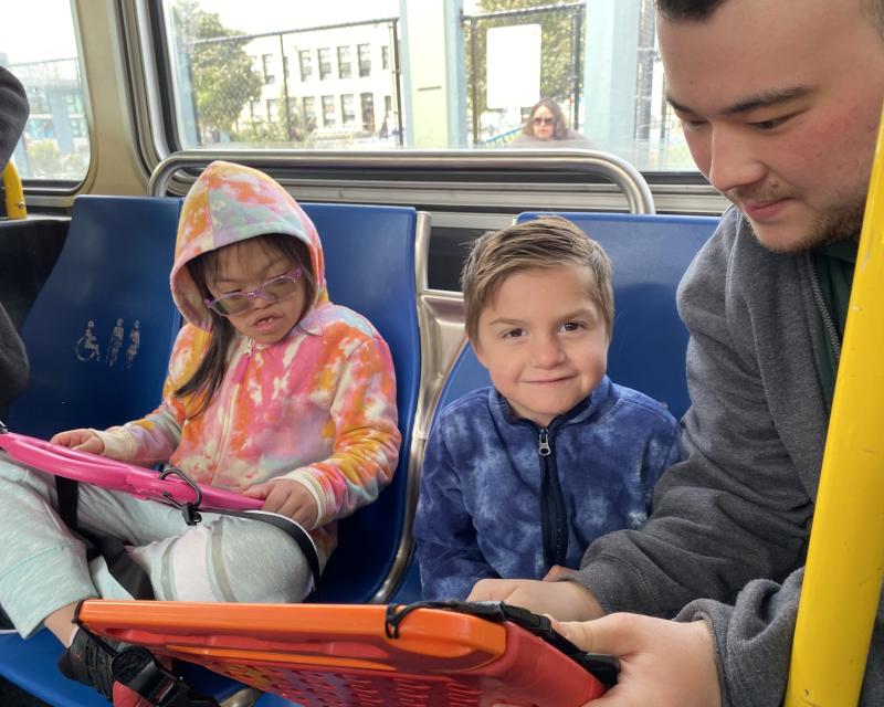 Two young children on a bus pay attention to an adult who shows them route information on a phone.