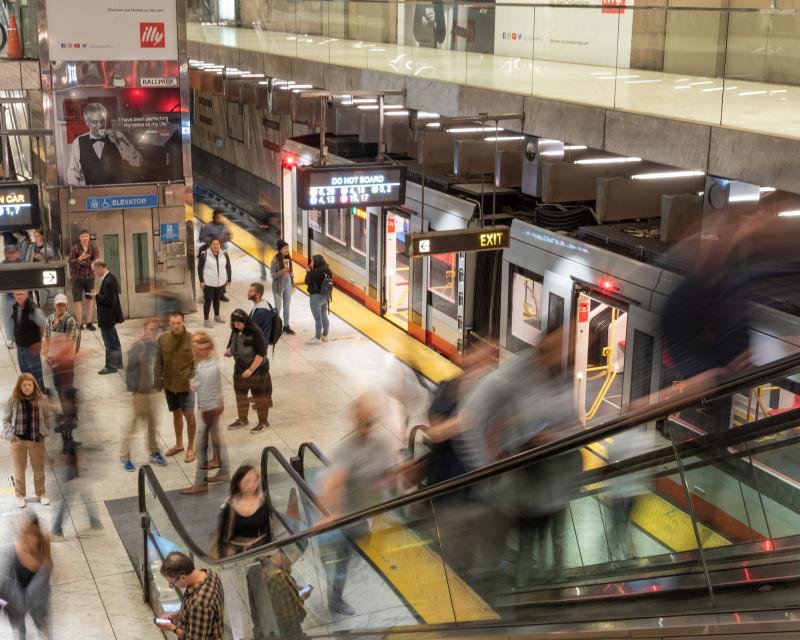 People wait for trains on a Muni platform as others exit on the escalator.