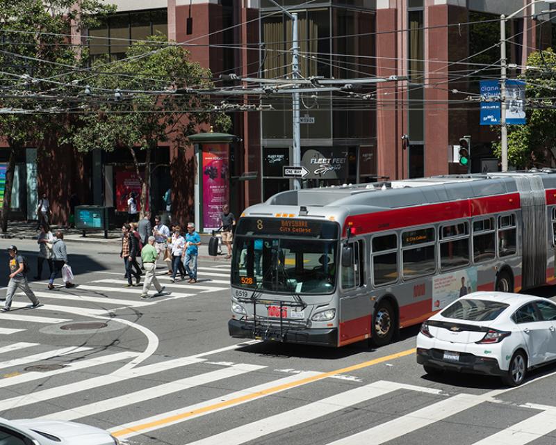 8 San Bruno bus crosses an intersection alongside people walking in the crosswalk.