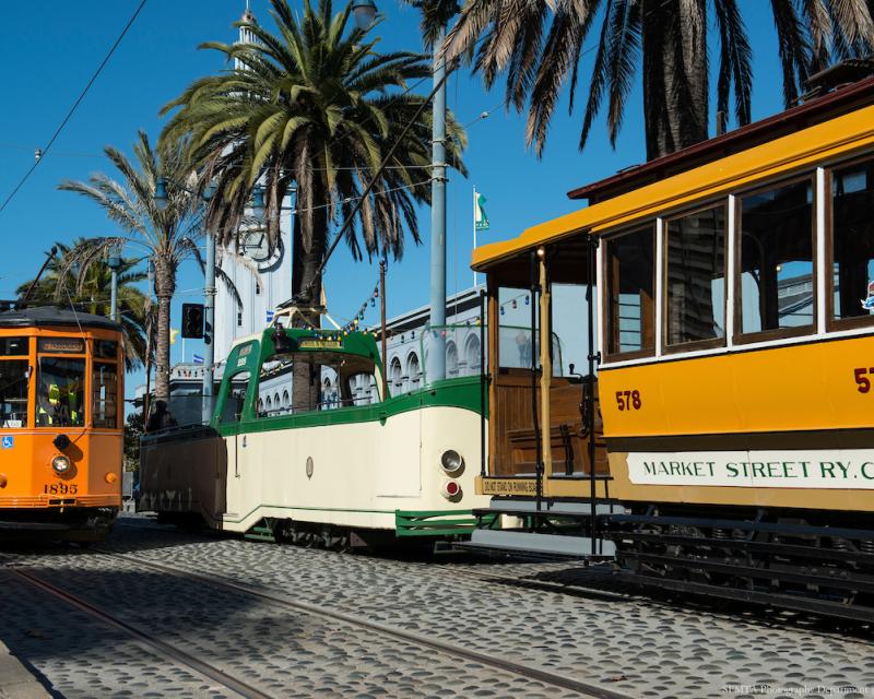 Three retro streetcars, including the Boat Tram, stop near palm trees by the Ferry Building.