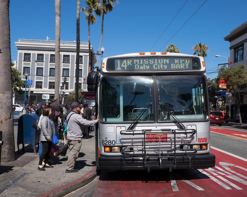 A photograph of people boarding the 14R Mission Rapid. Passengers board from a transit bulb outside the 16th Street BART station in the Mission District. The bus is in a red transit lane. 