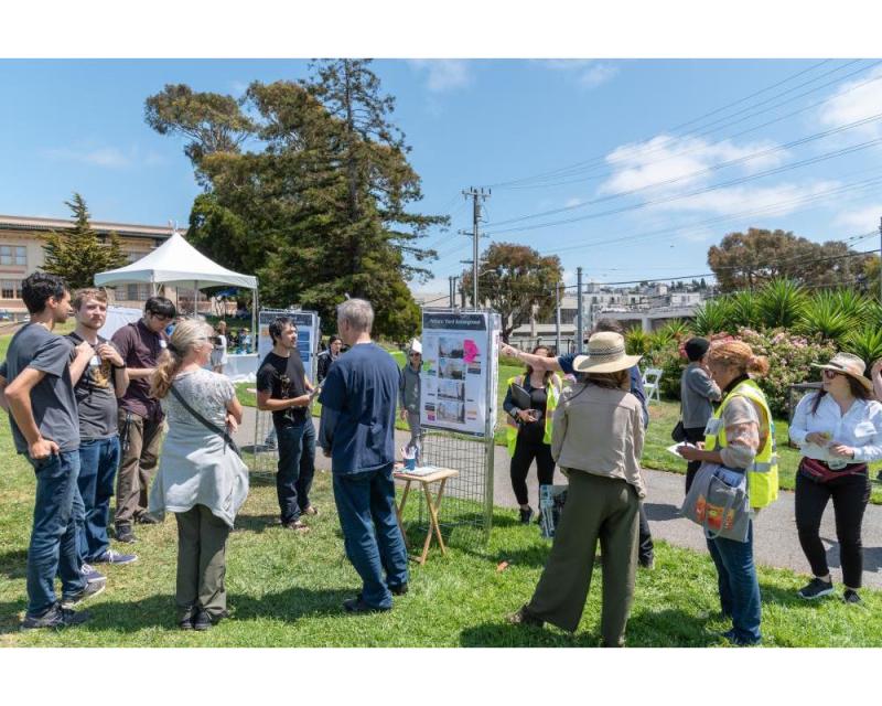 Image shows a group of people standing on a grassy hill, chatting near poster boards about Potrero Yard. 