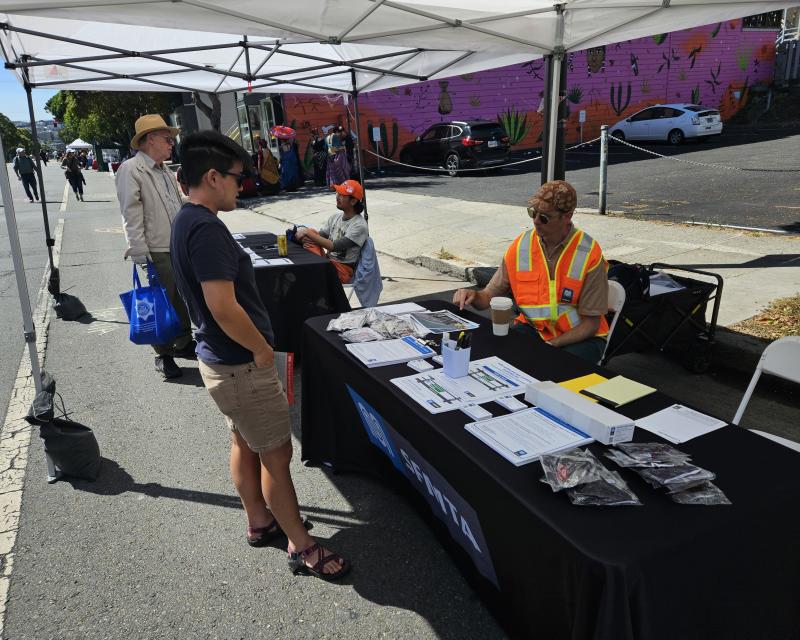 A member of the public is shown talking to an SFMTA staff person wearing an orange safety vest. A table, between them, is covered with diagrams and papers about the project.