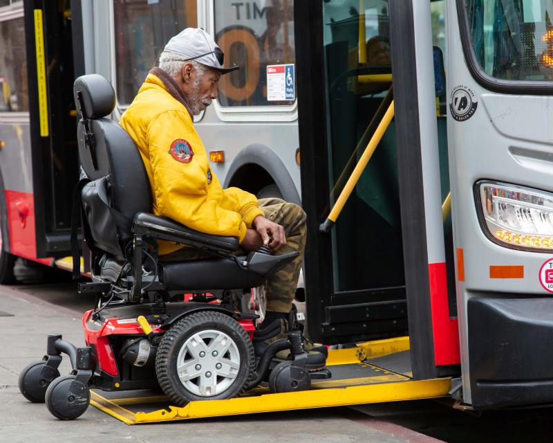 Person using a mobility device boards a Muni bus through a ramp lowered to sidewalk level.