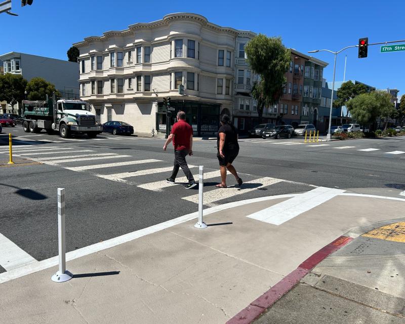 2 pedestrians crossing Guerrero at 17th, Yellow plastic posts at the center lines of the intersection and white plastic posts and khaki painted safety zone in the foreground at the corner of the intersection