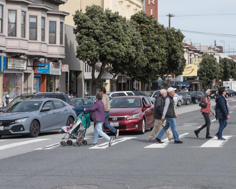 Several people cross Geary Boulevard as drivers behind the crosswalk. 