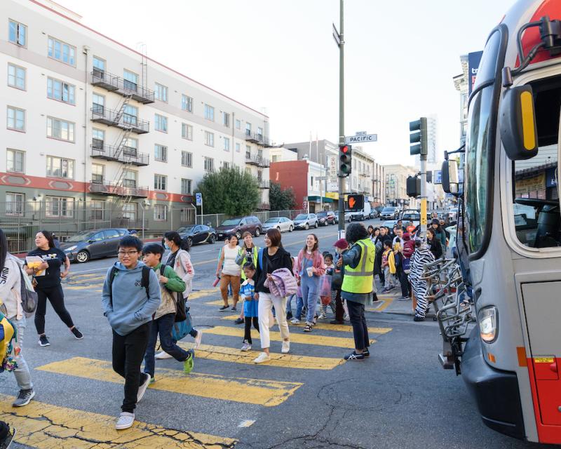 Students and families cross a street with help from a crossing guard. A bus pauses to let everyone through the intersecion.