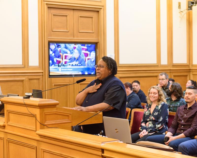 Denise Green holds speaks at a mic for the SFMTA Board of Directors special recognition award. A crowd sits in rows behind her.